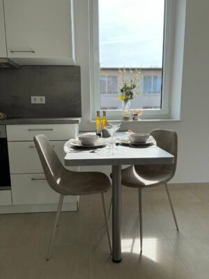 Moderne Küchen- und Esszimmergestaltung mit weißem Tisch, bequemen Stühlen, frischen Blumen und Sonnenschein durch das Fenster für ein einladendes Wohnerlebnis.