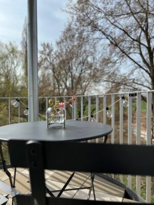 Gemütlicher Balkon mit schwarzmoderne Möbel, Blumenvase mit Kirschblüten, romantische Lichterkette, Blick ins Grüne, ideal für entspannte Momente.