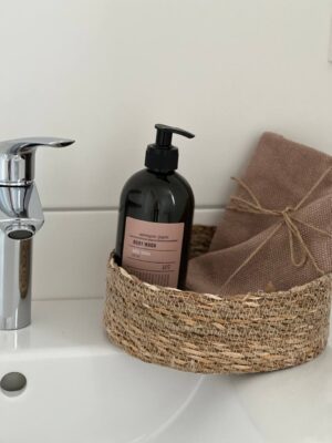 Flasche mit Body Wash und Handtuch im Korb auf Waschbecken, modernes Badezimmereinrichtung, Hygieneartikel, stilvolle Badezimmerdekoration, nachhaltige Organisation, elegante Badaccessoires, wellnessorientiertes Badezimmer, natürliche Home-Elemente, Funktionalität und Design, Augenmerk auf Körperpflege und Hygiene.