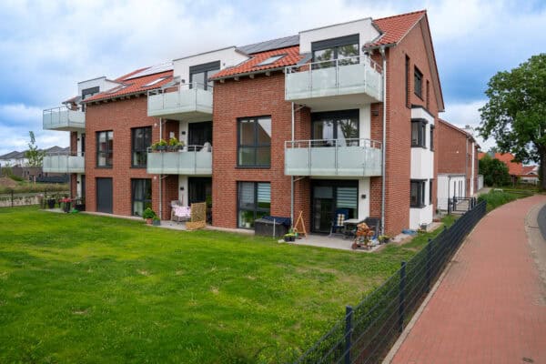 Moderner Wohnblock mit roten Ziegeln, weißen Fassaden und Balkonen, umgeben von grünem Garten und idyllischer Umgebung – ideal für zeitgemäßes Wohnen im Grünen.